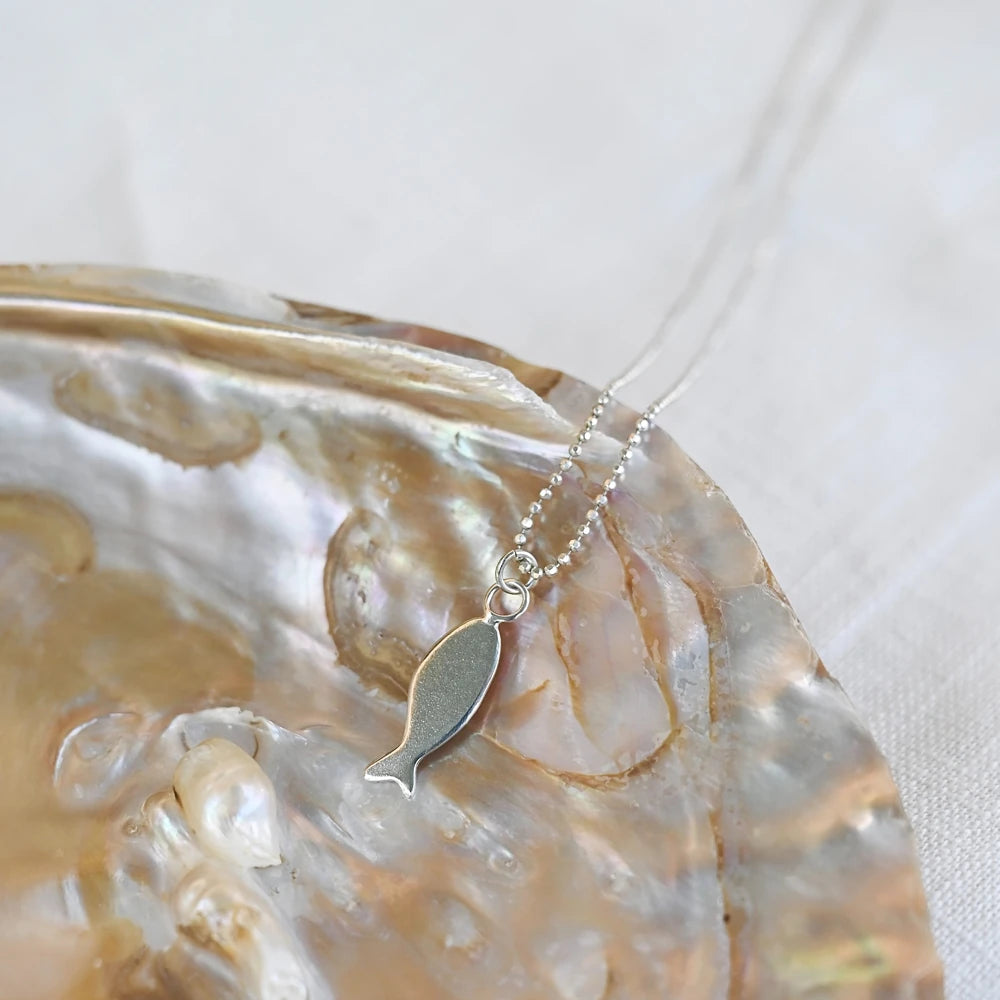 שרשרת דג כסף מונחת בתוך צדף פנינה מבריק, תכשיט אמירה מינימליסטי בהשראת עולם המים והים.