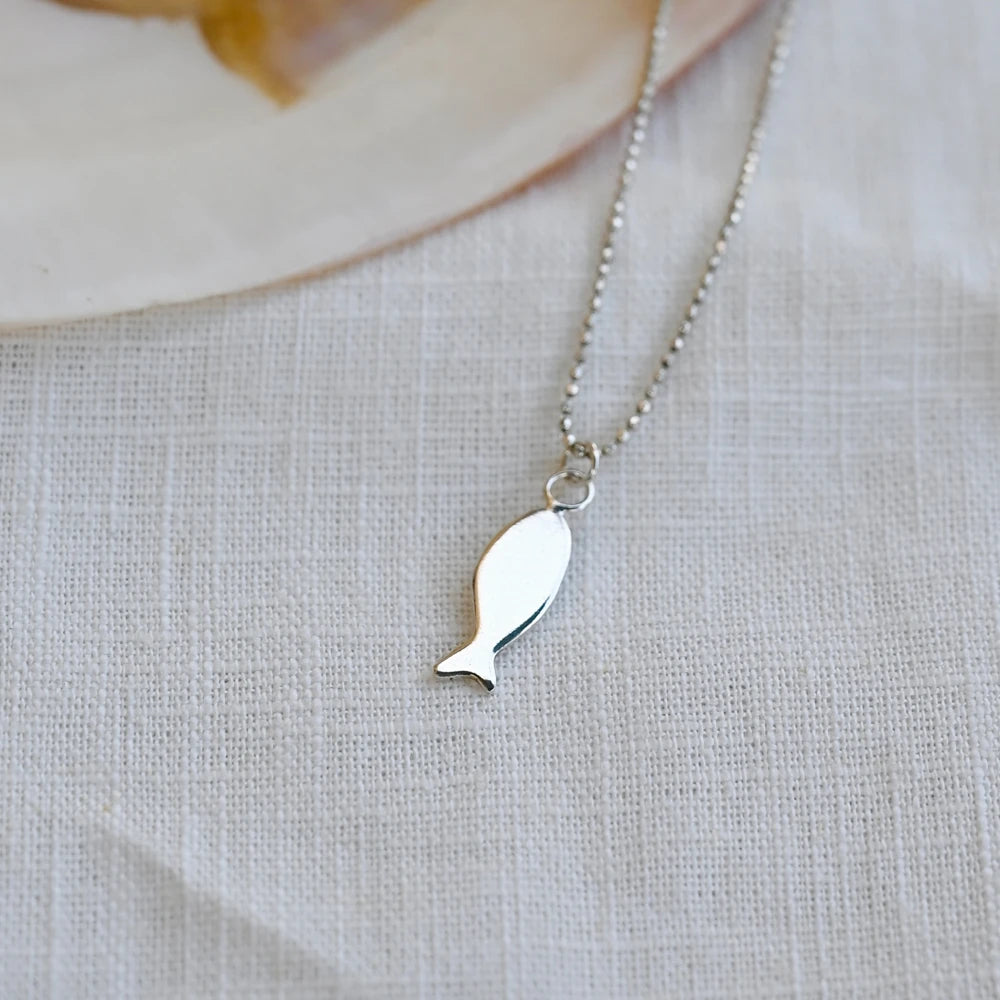 תליון דג מכסף סטרלינג על שרשרת עדינה, מבט חזיתי על רקע בד פשתן לבן המדגיש את הקווים הנקיים של התכשיט.