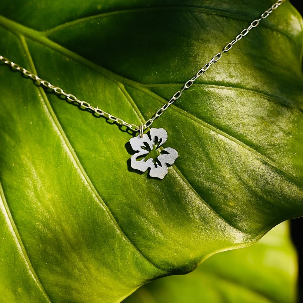 שרשרת כסף 925 עם תליון פרח היביסקוס בעיצוב פתוח ואורגני, מונחת על עלה ירוק בתאורה טבעית.
