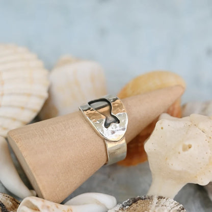 תקריב על טבעת דג מכסף בעיצוב פתוח ונוח, מונחת על מעמד עץ לצד צדפים טבעיים.