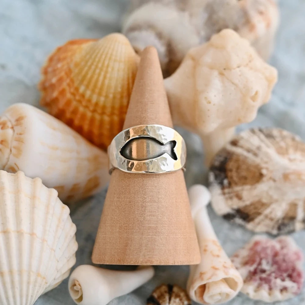 טבעת כסף 925 בעיצוב דג המזל על קונוס עץ, מוקפת צדפים באווירת ים וטבע.