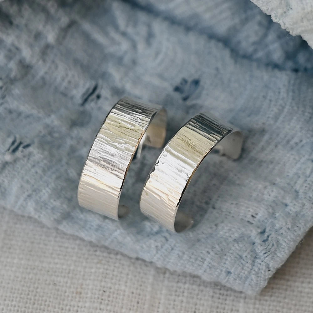 זוג עגילי חישוק מכסף סטרלינג עם טקסטורת פסים בעבודת יד, מונחים על בד כחול רך.