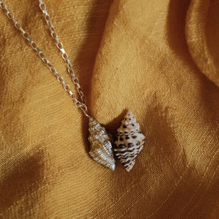 מבט מקרוב על שרשרת קונכייה מכסף 925 לצד קונכייה טבעית על רקע בד חום, המדגיש את הטקסטורה המפורטת.