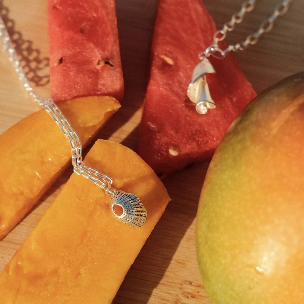 תליון צדף כסף בעיצוב 'חלון לים' מונח לצד פירות מנגו ואבטיח, משקף אווירת חופש וקיץ ישראלי.