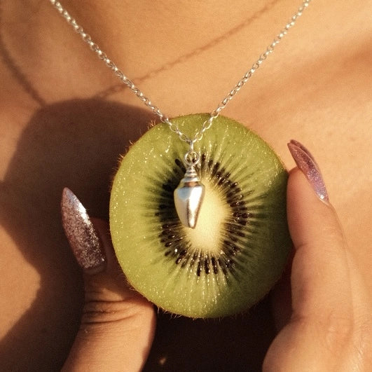 שרשרת כסף עם תליון קונכייה קטנה לצד פרי קיווי רענן, מדגישה את האופי הקיצי והצבעוני של קולקציית שירו.