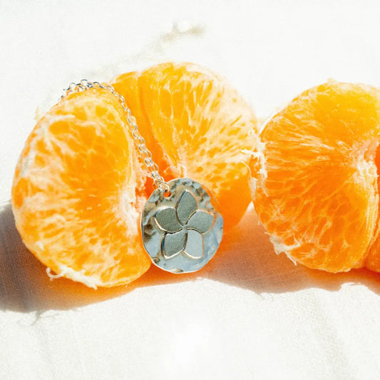 שרשרת כסף 925 עם תליון פרח פלומריה בעיצוב אורגני, מונחת על קלמנטינה רעננה כחלק מקולקציית הטבע של Shiroo.