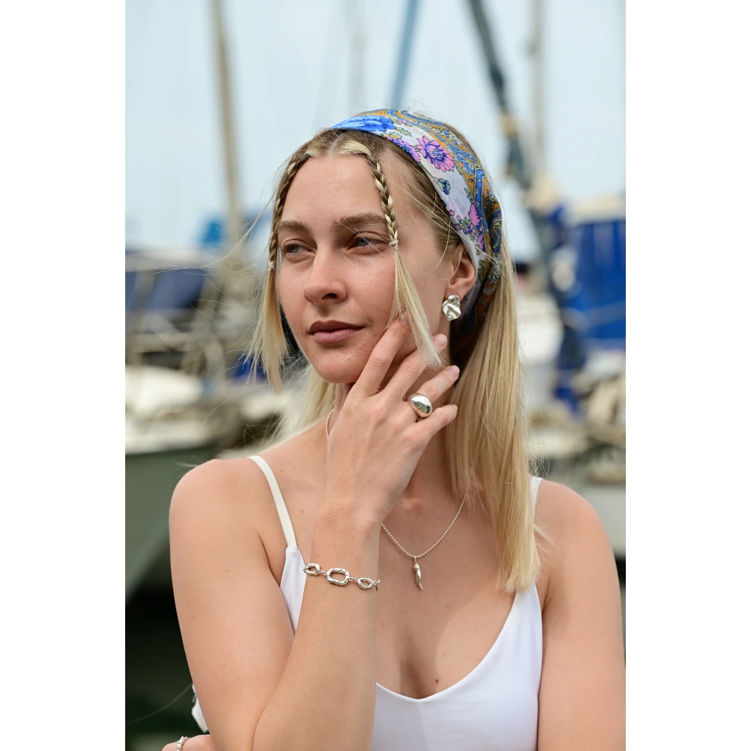 עגילי כסף יוליה של SHIROO, דוגמנית בלונדינית על רקע נמל. בסטיילינג עם שרשרת כסף עם תליון חרב, טבעת כסף גדולה וצמיד כסף חוליות. עגילי כסף סטרלינג 925 תוצרת ישראל.