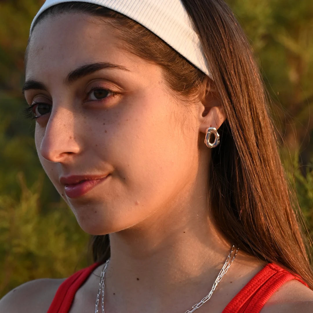 עגיל כסף לולאה חלול של SHIROO על אוזן דוגמנית ברונטית עם כובע לבן, על רקע טבע בשעת שקיעה. בשילוב שרשרת כסף חוליות. עגיל כסף 925 עבודת יד תוצרת ישראל.
