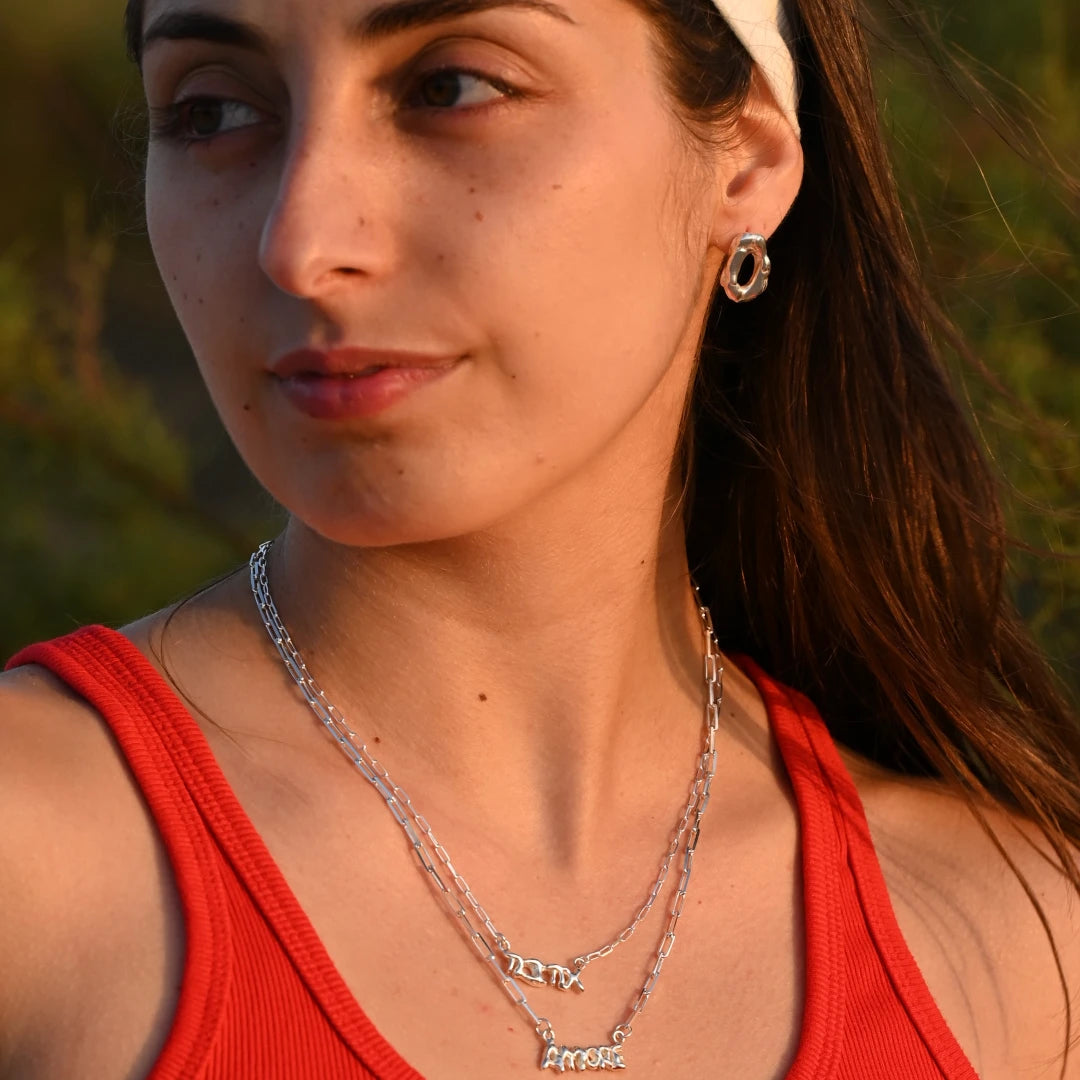 עגיל כסף לולאה חלול של SHIROO על אוזן דוגמנית ברונטית עם כובע לבן, בסטיילינג שכבות עם שרשרות כסף חוליות ושרשרת Amore. צילום על רקע טבע בשעת שקיעה. עגיל כסף 925 עבודת יד תוצרת ישראל.