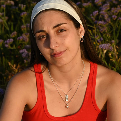 עגיל כסף חישוק ארוך של SHIROO על אוזן דוגמנית ברונטית עם כובע לבן, על רקע פרחים סגולים בשקיעה. בשילוב שרשרות כסף גלים לאורך. עגיל כסף 925 עבודת יד תוצרת ישראל.